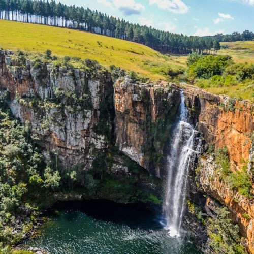A tall waterfall cascades down a rocky cliff into a pool, surrounded by lush greenery and a forest on top of the cliff. The sky is bright with scattered clouds, highlighting the vibrant landscape.