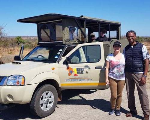 A man and woman stand smiling in front of a safari vehicle with a canopy. The background shows sparse vegetation and a clear sky, suggesting a warm, dry climate.