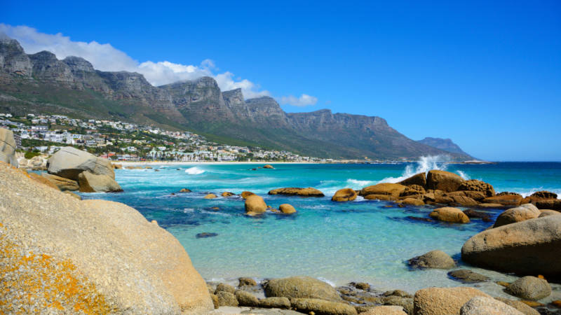 camps bay Experience the Best of the Cape Tour along a rocky shoreline with turquoise waters where waves crash against boulders. Rugged mountains rise in the distance under a clear blue sky, while sunlit, white buildings pepper the vibrant hillsides.