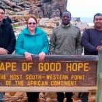 Four people stand behind a sign reading Cape of Good Hope, the most south-western point of the African continent. The sign, a highlight of the Cape Peninsula tour, is set against rocky terrain with the ocean visible in the background.