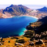 Experience a breathtaking view of a coastal landscape on our private cape peninsula tour, featuring a vibrant blue bay surrounded by rugged mountains. The shoreline is dotted with small towns and lush greenery, while the sun casts warm tones over the idyllic scene.