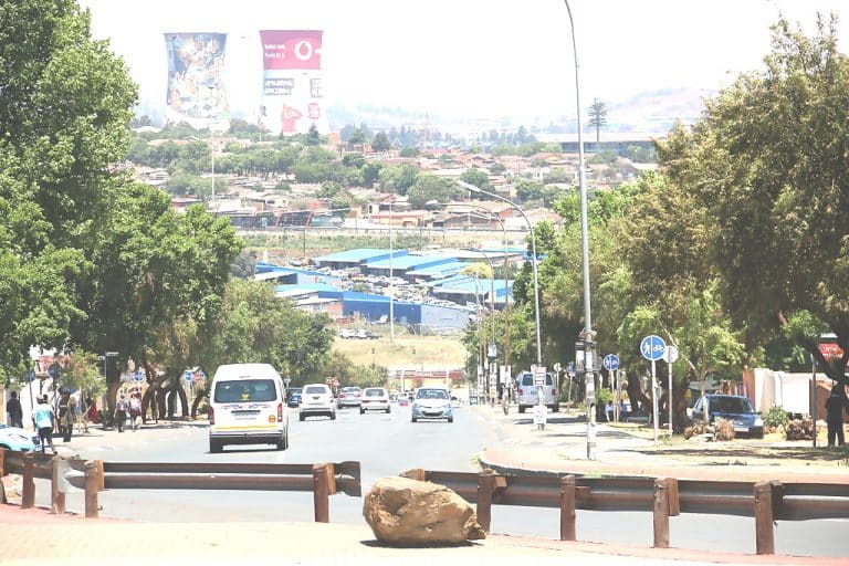 A busy urban street in South Africa lined with trees and streetlights. Vehicles, including a white minibus, travel along the road. In the background, residential areas and billboards are visible under a clear sky.