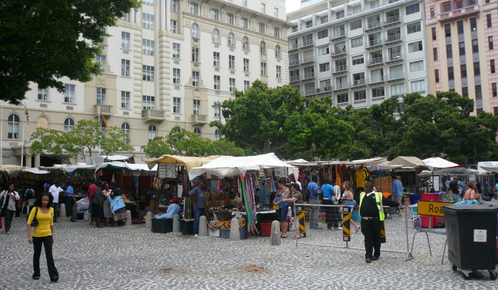 A lively outdoor market with stalls under white canopies, offering various goods. Shoppers walk along a cobblestone path. Surrounding the market are tall buildings and green trees, adding an urban backdrop to the bustling scene.