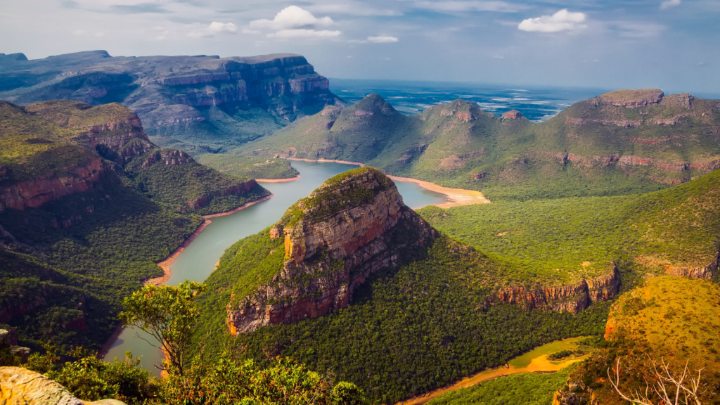 A stunning landscape featuring a winding river surrounded by lush green mountains and cliffs under a blue sky with scattered clouds. The view includes expansive valleys and diverse terrain.