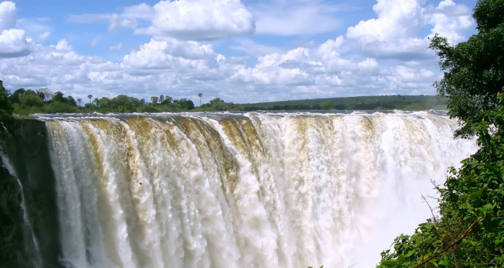 A powerful waterfall cascades over a wide cliff under a bright blue sky with scattered clouds. Lush green foliage surrounds the foreground, framing the view of the majestic waterfall.