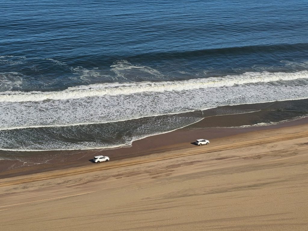 Two white cars drive along the sandy shoreline of a beach. The ocean waves gently crash nearby under a clear blue sky, creating a serene coastal scene. Tour Namibia today!