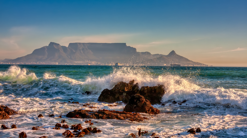 One of Cape Town attractions: a scenic view of Table Mountain with waves crashing against rocks, set against a clear blue sky and vibrant white-foamed sea.