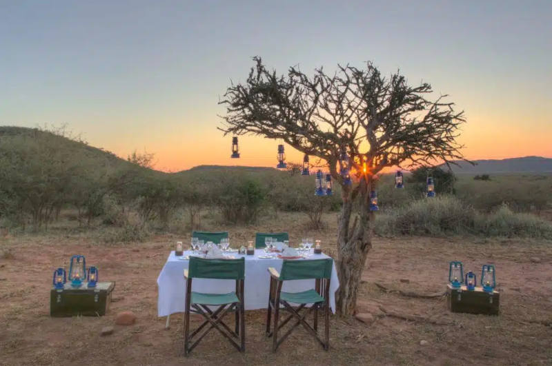 Madikwe Game Reserve serene outdoor dining setup at sunset in a savannah landscape. Four chairs surround a table with a white tablecloth and tableware. A tree decorated with lanterns stands nearby, with the sun setting in the background.